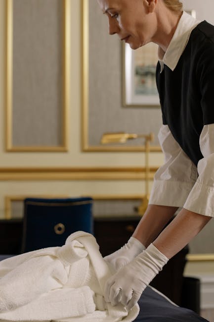 A female cleaner with light brown hair, wearing a yellow uniform and a blue lanyard, stands in a well-lit living room holding cleaning supplies. She has yellow rubber gloves on her hands and is holdin